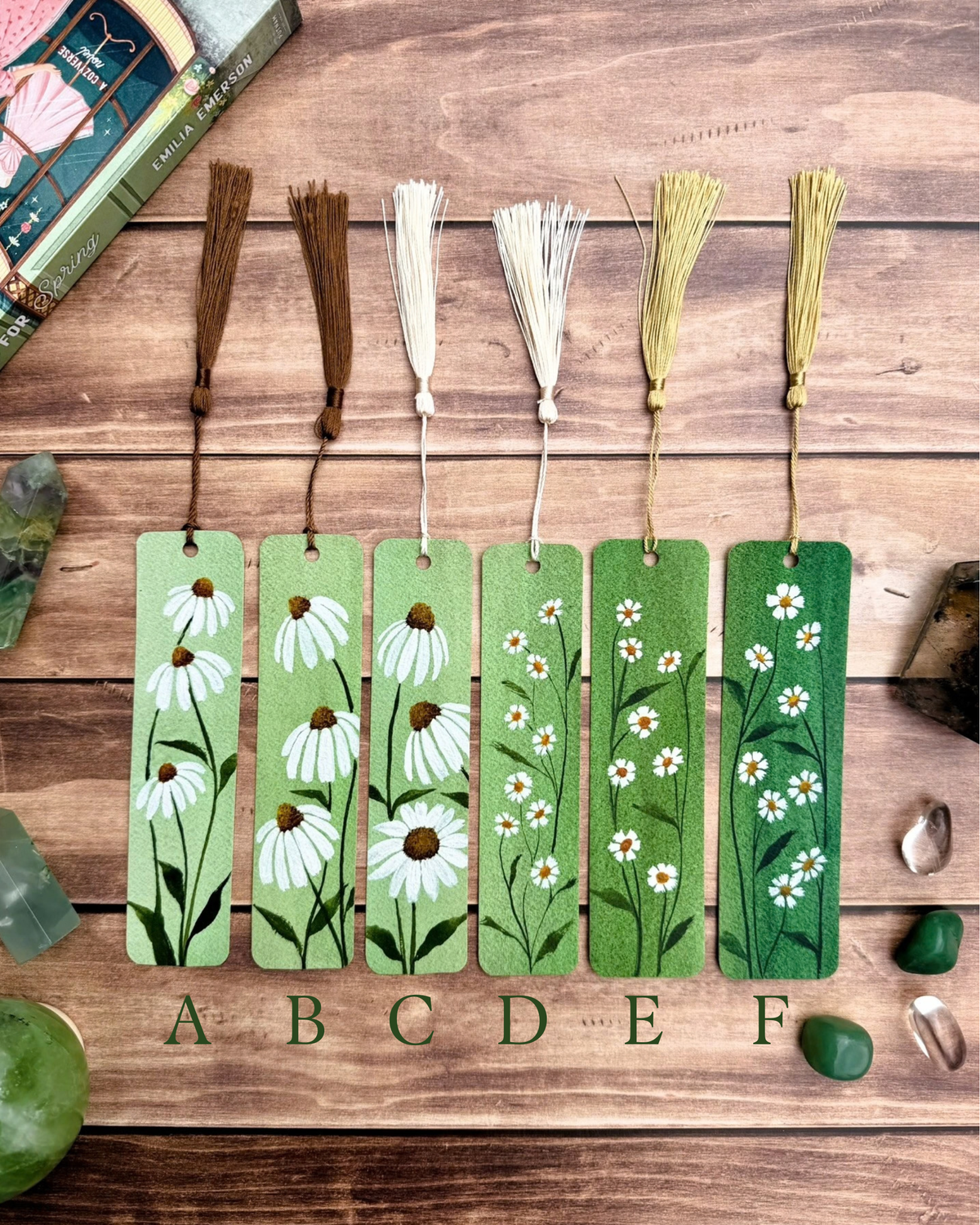 Six rectangular wooden bookmarks with tassel cords are arranged on a rustic wood surface. Each bookmark features a different floral illustration in white, green, and brown tones, labeled A through F below.  Echinacea and chamomile are painted on the bookmarks.