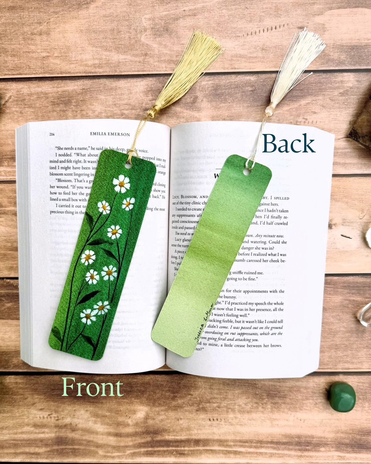 Two decorative bookmarks displayed on an open book on a wooden surface. One bookmark is a solid light green and reads 'Back', while the other is a green gradient with white chamomile floral patterns and a gold tassel, labeled 'Front'.
